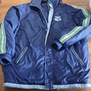 Op de afbeelding: Een marineblauwe jas met capuchon, lichtblauwe voering, knoopsluiting aan de voorzijde en lange mouwen met limoengroene en zwarte patroonstrepen aan de zijkanten. Op de borst staat het Diadora-logo in wit en blauw op een wit ovaal.