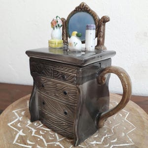 Può includere: Una teiera in ceramica a forma di cassettone con uno specchio e cassetti. La teiera è marrone con un motivo a venature del legno e ha un manico sul lato. La parte superiore della teiera ha un coperchio con uno specchio piccolo e alcuni oggetti decorativi in cima.
