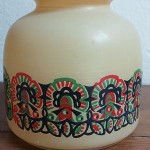Könnte beinhalten: Ein cremefarbenes Glasgefäß mit einem dekorativen Blumenband in Rot, Grün und Schwarz. Das Gefäß hat einen abgerundeten Körper und einen bauchigen Deckel, ideal zur Aufbewahrung oder Präsentation.