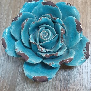 May include: A ceramic rose sculpture, turquoise in colour with distressed brown edges. The decorative flower is displayed on a wooden surface. The sculpture is approximately 20 cm in diameter.