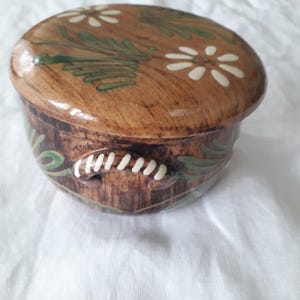 Può includere: Un contenitore in ceramica marrone con un coperchio, che presenta un design floreale dipinto a mano in verde e bianco. Il contenitore ha due maniglie sui lati.