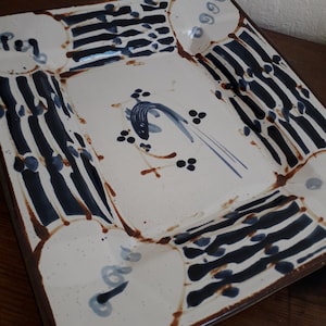 Può includere: Un vassoio da portata in ceramica bianca con un design a strisce blu e nere. Il vassoio ha un bordo rialzato e un design decorativo al centro.