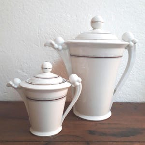 Peut inclure: Deux théières en porcelaine blanche avec des garnitures dorées et une bande brune. La plus grande théière a un couvercle et deux poignées. La plus petite théière a un couvercle et une seule poignée.