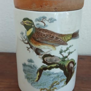 May include: A white ceramic jar with a wooden lid, decorated with a detailed illustration of a bird on a branch. The bird has brown, yellow, and green plumage. The jar has text at the base that reads "CURL BUNTING".