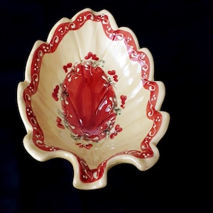 May include: A yellow ceramic bowl shaped like a leaf with red and white painted details. The bowl has a scalloped edge and a red floral design in the center.