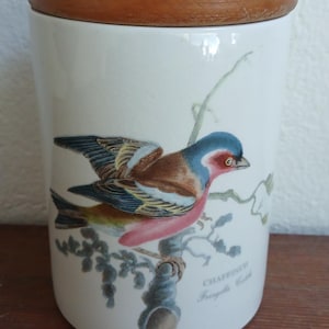 May include: A white ceramic jar with a wooden lid. The jar features a detailed illustration of a Chaffinch bird with blue, brown, and pink plumage perched on a branch. The text "CHAFFINCH" is below the bird.