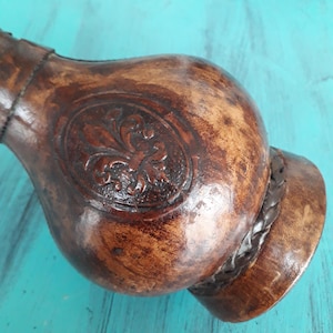 Könnte beinhalten: Eine braune Lederflasche mit einem dekorativen Blumenmuster. Die Flasche ist aus Leder und hat einen geflochtenen Lederriemen um den Hals.
