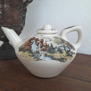 Może przedstawiać: Mały biały ceramiczny czajniczek z ręcznie malowanym motywem wodospadu i lasu. Czajniczek ma pokrywkę z brązowym uchwytem i brązowym dzióbkiem.