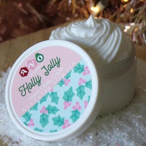 May include: A jar of white hand and body cream with a swirl design, next to its lid. The lid is white with a pink and blue design and the words "Holly Jolly" and "Avo Honey" printed on it. The background is a blurred Christmas tree.