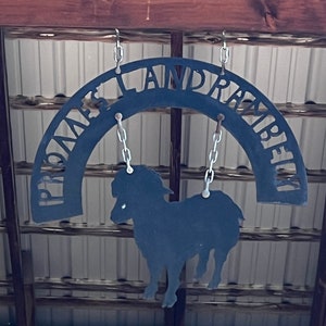 K&ouml;nnte beinhalten: Schwarzes Metallschild mit den Worten "Prince Island Ranch" in einer geschwungenen Bogenform. Eine Silhouette eines Schafes h&auml;ngt an dem Schild.