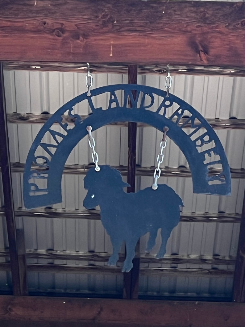 K&ouml;nnte beinhalten: Schwarzes Metallschild mit den Worten "Prince Island Ranch" in einer geschwungenen Bogenform. Eine Silhouette eines Schafes h&auml;ngt an dem Schild.