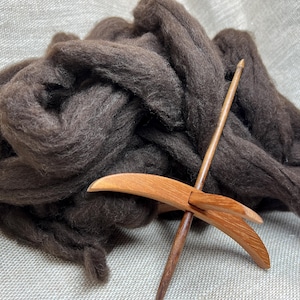 May include: A wooden drop spindle with a curved whorl positioned in front of a large, loose pile of dark brown unspun wool roving. The spindle features a slender, pointed shaft and a smooth, polished finish on the light brown wood grain.