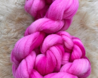 Könnte beinhalten: Ein leuchtend rosa Kammzuggarn, fest geflochten, mit verschiedenen Rosatönen. Das Garn wird auf einer hellbeige, strukturierten Oberfläche präsentiert, wodurch ein Kontrast von Farben und Texturen entsteht. Dieses Faserkunstmaterial ist ideal für verschiedene Bastelprojekte.