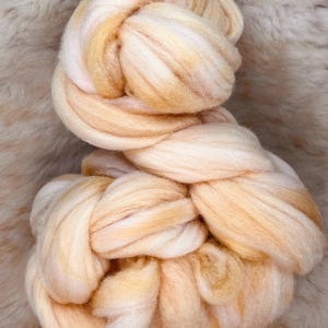 May include: A bundle of soft, white and peach colored wool roving. The roving is twisted and knotted together, creating a textured and fluffy appearance.