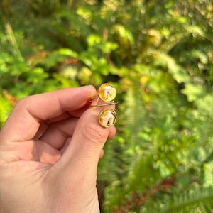 May include: A gold-toned ring featuring two circular settings, each containing a small, white, tooth-shaped element encased in a clear, amber-colored resin. The ring has a thin, open-style band that wraps around the finger.
