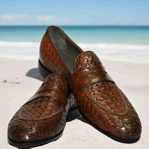 May include: A pair of brown leather loafers with a woven pattern. The loafers are on a white sandy beach.