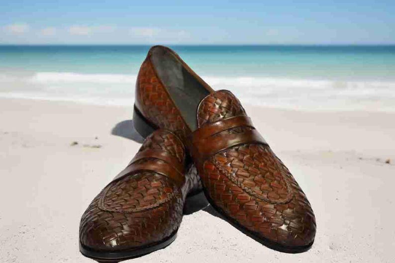 May include: A pair of brown leather loafers with a woven pattern. The loafers are on a white sandy beach.