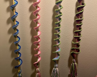 May include: Four handmade crochet spiral decorations in blue, pink, green, and purple. Each spiral has a tassel at the bottom. The spirals are hanging against a neutral wall. The crochet work is detailed and colorful.