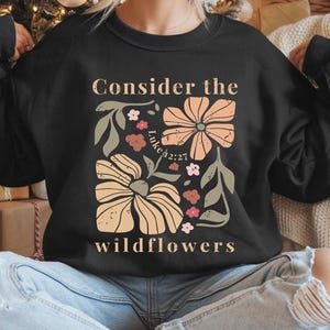 Puede incluir: Sudadera negra con un dise&ntilde;o floral y el texto "Consider the wildflowers" y "Luke 42:27".