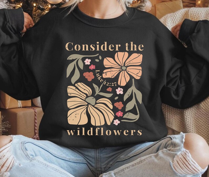 Puede incluir: Sudadera negra con un dise&ntilde;o floral y el texto "Consider the wildflowers" y "Luke 42:27".