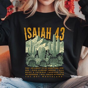 Puede incluir: Sudadera negra con un estampado gr&aacute;fico de un paisaje des&eacute;rtico con cactus y el texto "ISAIAH 43". El texto debajo del gr&aacute;fico dice: "FOR I AM ABOUT TO DO SOMETHING NEW. SEE, I HAVE ALREADY BEGUN! I WILL MAKE A PATHWAY THROUGH THE WILDERNESS, I WILL CREATE RIVERS IN THE DRY WASTELAND."