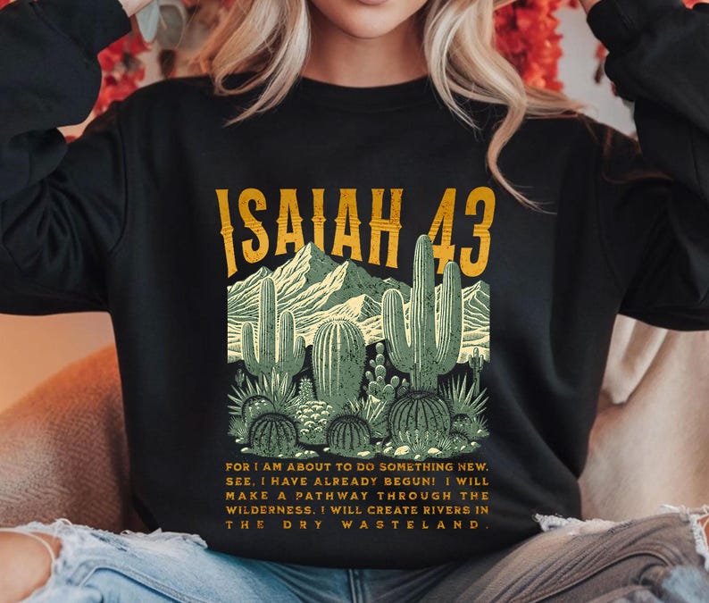 Puede incluir: Sudadera negra con un estampado gr&aacute;fico de un paisaje des&eacute;rtico con cactus y el texto "ISAIAH 43". El texto debajo del gr&aacute;fico dice: "FOR I AM ABOUT TO DO SOMETHING NEW. SEE, I HAVE ALREADY BEGUN! I WILL MAKE A PATHWAY THROUGH THE WILDERNESS, I WILL CREATE RIVERS IN THE DRY WASTELAND."