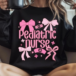 以下が含まれることがあります： ピンクのリボンと「Pediatric + Nurse +」の文字がプリントされた黒いスウェットシャツ