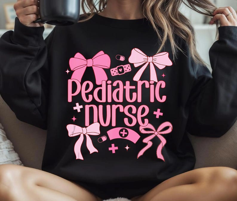 以下が含まれることがあります： ピンクのリボンと「Pediatric + Nurse +」の文字がプリントされた黒いスウェットシャツ