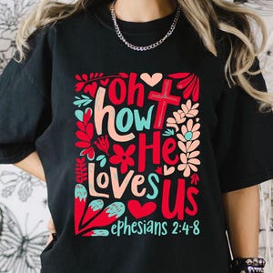 Puede incluir: Camiseta negra con un dise&ntilde;o gr&aacute;fico colorido que dice "Oh how He loves us" con una cruz y flores. El texto "Efesios 2:4-8" tambi&eacute;n est&aacute; incluido.