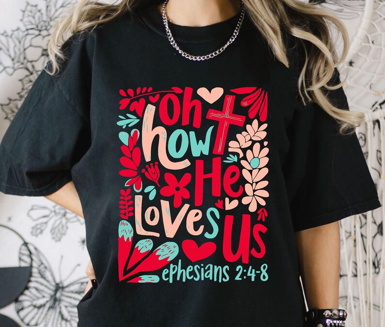 Puede incluir: Camiseta negra con un dise&ntilde;o gr&aacute;fico colorido que dice "Oh how He loves us" con una cruz y flores. El texto "Efesios 2:4-8" tambi&eacute;n est&aacute; incluido.