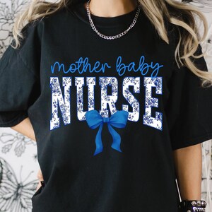 以下が含まれることがあります： 白い花柄と青いリボンが付いた黒いTシャツ。Tシャツには「mother baby NURSE」の文字がプリントされています。