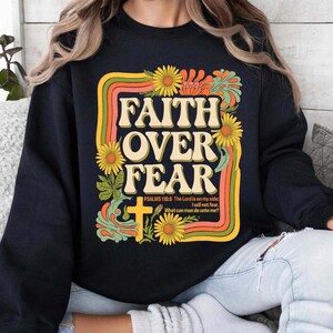 Może przedstawiać: Czarna bluza z nadrukiem w stylu retro z napisem "FAITH OVER FEAR" w ż&oacute;łtych i pomarańczowych literach. Grafika zawiera r&oacute;wnież krzyż, kwiaty i werset biblijny "PSALMS 118:6 The Lord is on my side I will not fear. What can man do unto me?"