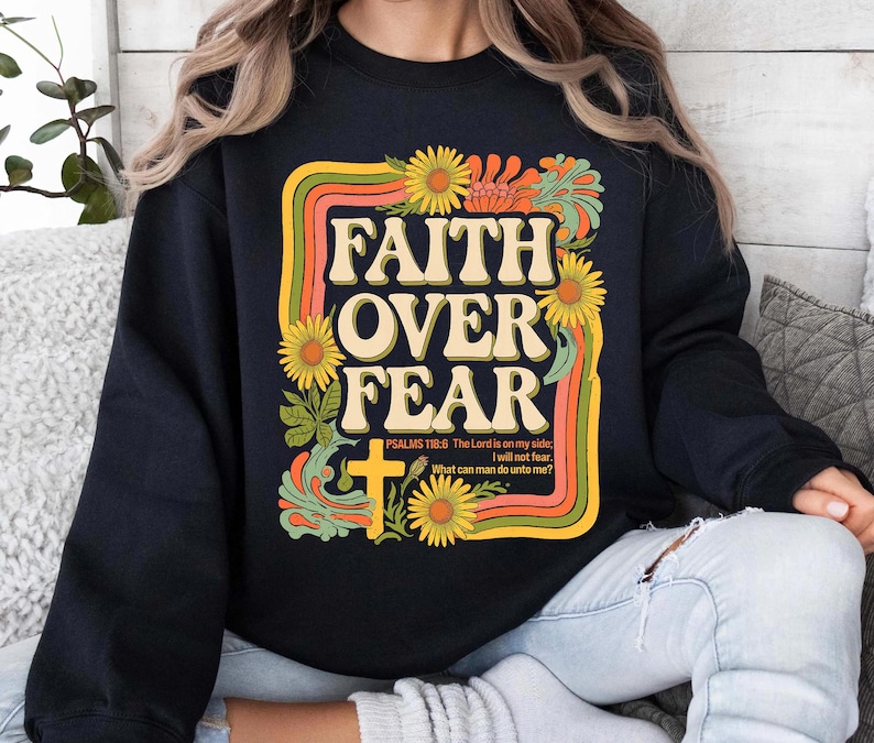 Może przedstawiać: Czarna bluza z nadrukiem w stylu retro z napisem "FAITH OVER FEAR" w ż&oacute;łtych i pomarańczowych literach. Grafika zawiera r&oacute;wnież krzyż, kwiaty i werset biblijny "PSALMS 118:6 The Lord is on my side I will not fear. What can man do unto me?"
