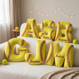 May include: Yellow softball-themed letter pillows with red stitching and personalized names: Asher, Sophia, Benjamin, Grayson, Josephine, and Madison. Softballs are also visible. Decorative and personalized.
