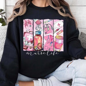 以下が含まれることがあります： 医療用品と「#nurselife」というテキストがピンクと白でグラフィックプリントされた黒いスウェットシャツ。
