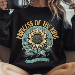 Puede incluir: Sudadera negra con un gr&aacute;fico vintage de un girasol y el texto "Princess of the King Ephesians 1:5".