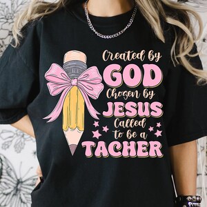 Może przedstawiać: Czarny T-shirt z r&oacute;żowym tekstem, kt&oacute;ry brzmi "Stworzony przez BOGA Wybrany przez JEZUSA Powołany, aby być NAUCZYCIELEM" i r&oacute;żową kokardką z ż&oacute;łtym oł&oacute;wkiem w środku.