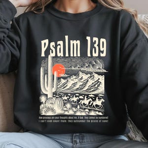 Puede incluir: Sudadera negra con un estampado gr&aacute;fico de un paisaje des&eacute;rtico, un cactus y el texto "Salmo 139". El texto debajo del gr&aacute;fico dice: "&iexcl;Cu&aacute;n preciosos son tus pensamientos acerca de m&iacute;, oh Dios! No se pueden contar. Ni siquiera puedo contarlos: &iexcl;superan en n&uacute;mero los granos de arena!"