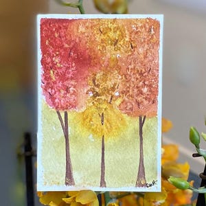 Könnte beinhalten: Aquarellmalerei von drei Bäumen mit braunen Stämmen und leuchtendem Herbstlaub in Rot-, Orange- und Gelbtönen. Der Hintergrund ist hellgrün. Das Kunstwerk ist unten rechts signiert.
