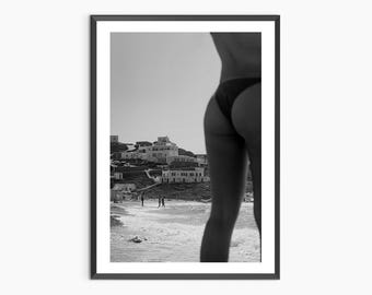 May include: Black and white photograph of a woman in a bikini standing on a beach with a small town in the background.