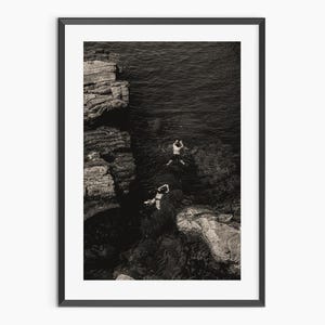 Peut inclure: Une photographie en noir et blanc de deux personnes sautant d'une falaise rocheuse dans l'eau. L'eau est sombre et les rochers sont rugueux et texturés.