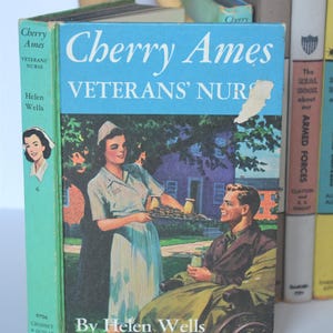 May include: Vintage book titled "Cherry Ames Veterans' Nurse" by Helen Wells. The cover features a nurse serving a tray to a man in a wheelchair. The book is light blue with white text and a colorful illustration. Other books are visible in the background.
