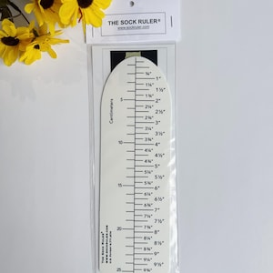 Puede incluir: Una regla para calcetines blanca con marcas y texto en negro, que mide hasta 24 cm. La regla también tiene medidas en pulgadas hasta 9 1/2 pulgadas. El texto "THE SOCK RULER" y la dirección del sitio web están impresos.