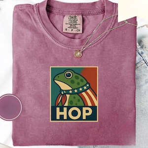 Puede incluir: Una camiseta Comfort Colors malva con un gráfico de rana y la palabra "HOP". La rana lleva un cuello patriótico y una bufanda a rayas rojas, blancas y azules. Un collar dorado con un colgante descansa sobre la camiseta.