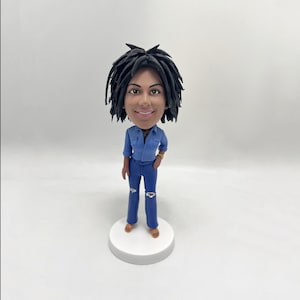 May include: A custom bobblehead figurine of a person with black hair and a blue denim shirt and jeans. The figurine is standing on a white base.