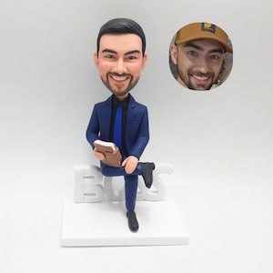 May include: A custom bobblehead figurine of a man in a blue suit, holding a brown notebook, seated on a white "BOSS" sign. A photo of a man in a brown cap is in the background. The figurine is on a white base.