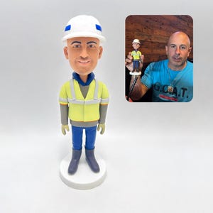 Może przedstawiać: Niestandardowa figurka z kiwającą głową mężczyzny w białym kasku, żółtej kamizelce odblaskowej, niebieskich dżinsach i szarych butach. Figurka stoi na białej podstawie. Mniejszą wersję trzyma mężczyzna w niebieskiej koszuli.