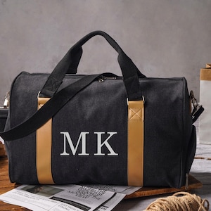 May include: A black duffel bag featuring two tan vertical accent strips and the white letters MK printed on the front. The bag includes black fabric handles and a matching shoulder strap, with a zipper closure along the top edge of the main compartment.