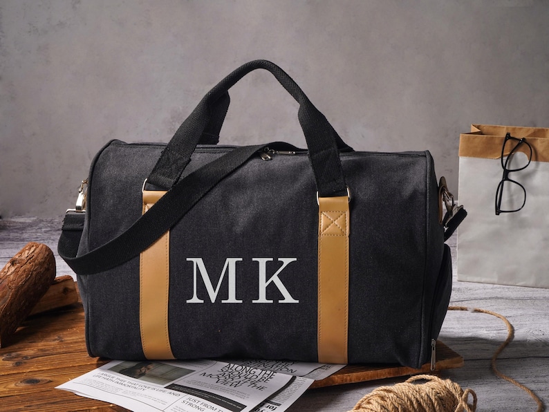 May include: A black duffel bag featuring two tan vertical accent strips and the white letters MK printed on the front. The bag includes black fabric handles and a matching shoulder strap, with a zipper closure along the top edge of the main compartment.