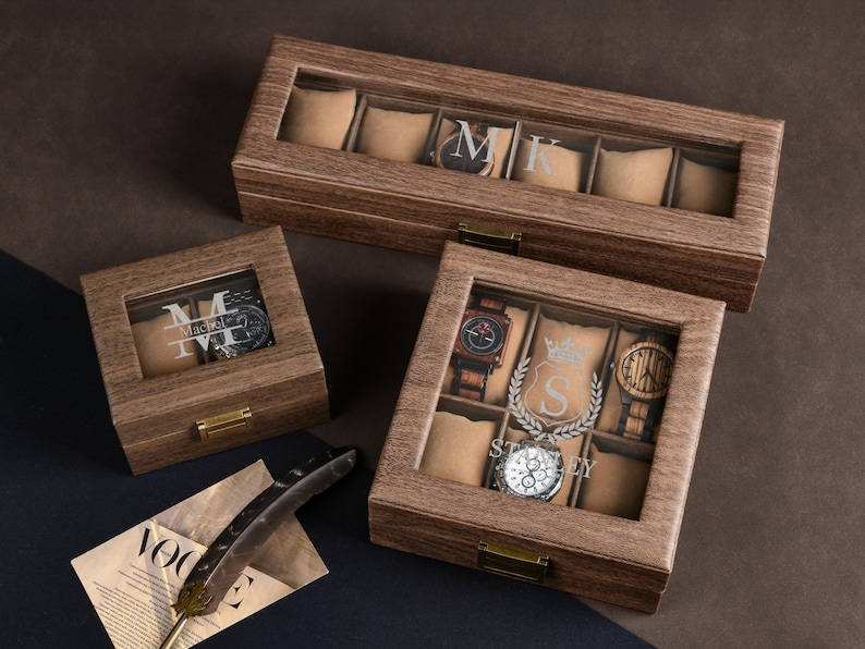 May include: Three wooden watch boxes with clear glass tops. The boxes are in different sizes and have individual watch compartments with beige cushions. The largest box has the initials "MK" etched on the glass. The smallest box has the name "Mackel" etched on the glass.